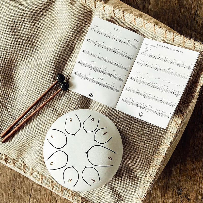 8 Tone 6-inch Steel Tongue Hank Tank Panda Drum White with Drumsticks and Musical Score Sheet on top of a white seat